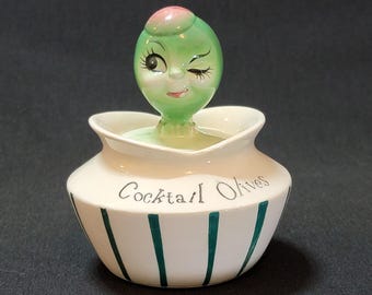 May include: A white ceramic olive dish with green stripes and the words "Cocktail Olives" on the side. The lid is a green olive with a winking face and a pink top.