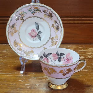 Puede incluir: Un juego de taza y platillo de té rosa y dorado con un diseño floral. La taza y el platillo presentan un delicado patrón de rosas rosas y blancas con detalles dorados.