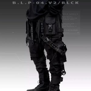 Può includere: Pantaloni cargo neri con più tasche, cinghie e fibbie. I pantaloni presentano le scritte "HOLY GRAIL" e "GREED" sulle cinghie. Il testo "B.L.P-04.V2/BLCK" è in alto. Stile streetwear o techwear.