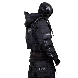 Puede incluir: Chaqueta y pantal&oacute;n t&aacute;cticos negros con m&uacute;ltiples bolsillos y correas. La chaqueta presenta el texto "H-G" en el pecho y el brazo, y "HOLYVERSE" en la correa del hombro. La persona lleva un casco negro.