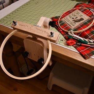 May include: A wooden embroidery hoop stand with a large hoop attached. The stand has a clamp to secure it to a table. A red and green plaid fabric with a small embroidery hoop is on the table. The embroidery hoop has a design that says "'Twill be Home for Christmas".