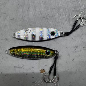 May include: Two fishing lures. The top lure is white with black and iridescent stripes. The bottom lure is black and gold with iridescent accents. Each lure has a hook and a black cord.
