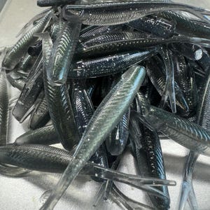 May include: A pile of black and green soft plastic fishing lures. The lures are shaped like fish and have a shimmering finish.