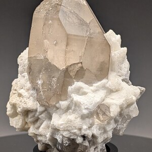 May include: A large, clear quartz crystal with a smoky brown hue, perched atop a bed of white calcite crystals. The crystal is displayed on a black base.