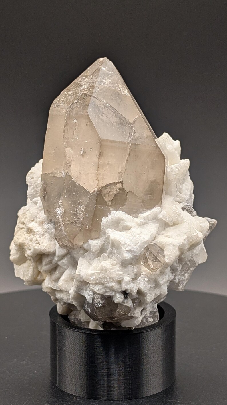 May include: A large, clear quartz crystal with a smoky brown hue, perched atop a bed of white calcite crystals. The crystal is displayed on a black base.