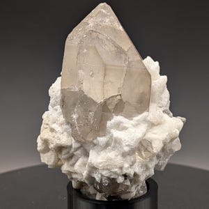 May include: A large, clear quartz crystal with a smoky brown hue sits atop a cluster of white, opaque crystals. The quartz crystal is faceted and has a pointed tip. The cluster of crystals is on a black base.