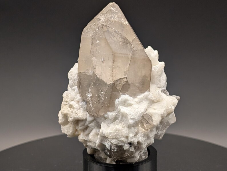 May include: A large, clear quartz crystal with a smoky brown hue sits atop a cluster of white, opaque crystals. The quartz crystal is faceted and has a pointed tip. The cluster of crystals is on a black base.