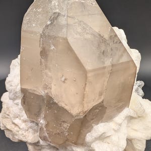 May include: A large, clear quartz crystal with a smoky brown hue, resting on a bed of white, crystalline matrix. The crystal is sharply pointed and has a faceted, geometric shape.