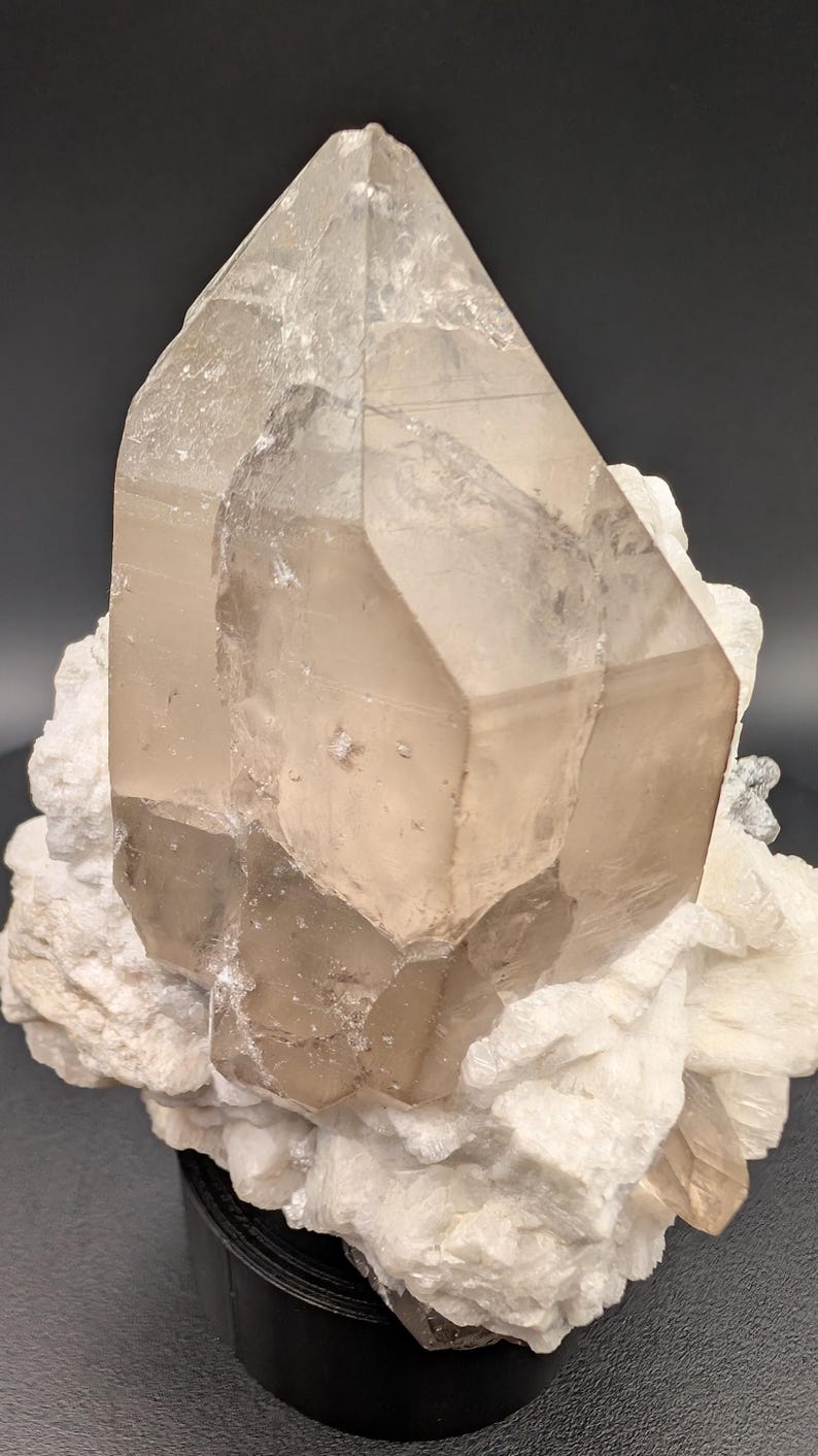 May include: A large, clear quartz crystal with a smoky brown hue, resting on a bed of white, crystalline matrix. The crystal is sharply pointed and has a faceted, geometric shape.