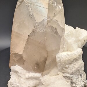 May include: A large, clear quartz crystal with a smoky brown hue sits atop a bed of white, crystallized rock. The crystal is on a black base.
