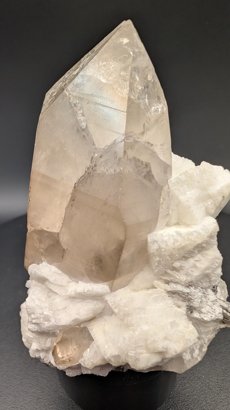 May include: A large, clear quartz crystal with a smoky brown hue sits atop a bed of white, crystallized rock. The crystal is on a black base.
