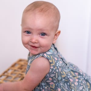 May include: A baby girl with blue eyes is wearing a sleeveless dress with a floral pattern in shades of blue, pink, and yellow. She is smiling and looking at the camera.