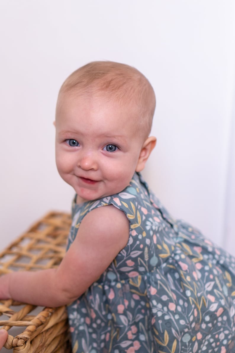 May include: A baby girl with blue eyes is wearing a sleeveless dress with a floral pattern in shades of blue, pink, and yellow. She is smiling and looking at the camera.