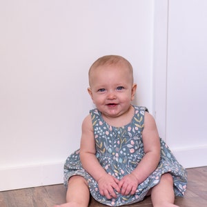 May include: A baby girl wearing a light blue dress with a floral pattern sits on a wooden floor in front of a white wall.