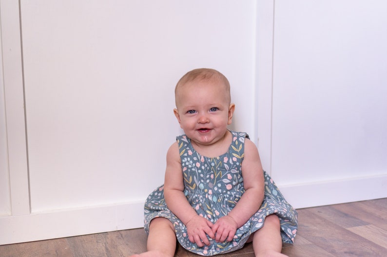 May include: A baby girl wearing a light blue dress with a floral pattern sits on a wooden floor in front of a white wall.