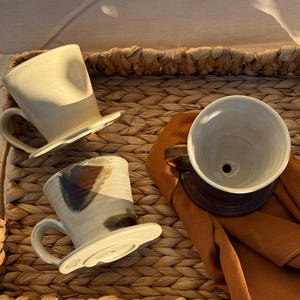 Könnte beinhalten: Drei Keramik-Kaffee-Pour-Over-Sets und eine Kaffeetasse liegen auf einem gewebten Tablett. Die Pour-Over-Sets sind cremefarben und weiß mit einem dunkelbraunen Sockel. Eine Tasse hat ein einzigartiges braunes und blaues Design. Ein braunes Tuch ist in der Nähe drapiert.