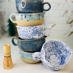 May include: A stack of five ceramic bowls with blue and white speckled glaze. The bowls have a handle on one side and are stacked in a graduated size. A bamboo whisk is in the foreground.