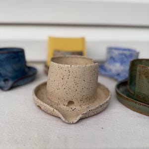 以下が含まれることがあります： ユニークなデザインの手作り陶器のコレクション。中央の作品は、広い縁と小さな注ぎ口が付いたベージュの斑点のある陶器です。他の作品には、同様の形状の青、緑、黄色の陶器が含まれています。