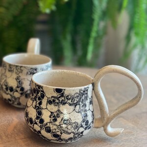 May include: Two ceramic mugs with a speckled, off-white glaze. The mugs are decorated with a repeating pattern of black skull illustrations. Each mug has a large, curved handle. Green foliage is visible in the background.