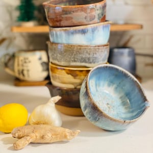 Peut inclure: Une pile de bols en céramique faits à la main de différentes couleurs, dont le marron, le bleu et le beige. Un citron, une tête d'ail et un morceau de gingembre sont au premier plan. Une tasse et un petit arbre décoratif sont en arrière-plan.