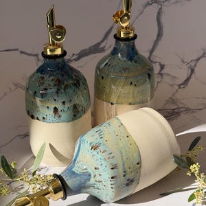 Op de afbeelding: Drie keramische oliedispensers met goudkleurige metalen schenktuiten. Elke fles heeft een tweekleurig ontwerp met een matte crèmekleurige basis en een glanzend, gespikkeld blauw en groen glazuur op het bovenste gedeelte.