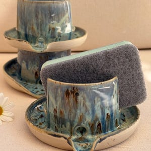 Può includere: Due porta spugne in ceramica con finitura smaltata a goccia blu, marrone e crema. Ogni supporto presenta una base circolare con un piccolo scarico e una fessura verticale per tenere in posizione verticale una spugna abrasiva grigia.