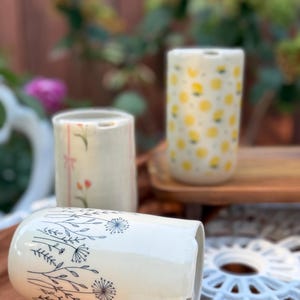 May include: Three cylindrical ceramic tumblers with distinct patterns. One features blue line-art wildflowers, another has a vertical pink stripe with small floral accents, and the third is decorated with a repeating yellow lemon print.