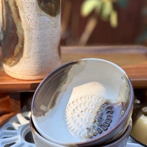 May include: A ceramic bowl with a cream-colored interior featuring a raised, circular, petal-like texture at the base, accented with brown glaze drips. The exterior is a smooth, light cream color with a dark brown rim and base.