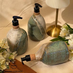 May include: Three handcrafted ceramic soap dispensers with black pumps and cork collars. The dispensers have a speckled, glazed finish in shades of green, white, and brown. The text "ADORE CERAMIC" and "100% Handcrafted" is visible.