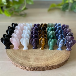 May include: A selection of small angel figurines in various colours, including black, pink, white, purple, green, and blue. The figurines are arranged on a round, wooden display. The background includes greenery.