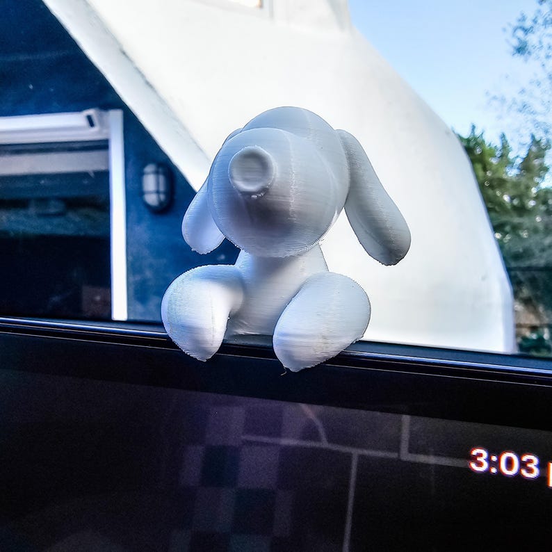 K&ouml;nnte beinhalten: Eine wei&szlig;es, 3D-gedrucktes Hundefigur mit gro&szlig;en Ohren und rundem K&ouml;rper. Der Hund sitzt auf einer dunklen Oberfl&auml;che, m&ouml;glicherweise einem Armaturenbrett, mit einem Fenster und der Au&szlig;enansicht im Hintergrund. Die Uhrzeit 15:03 Uhr wird angezeigt.