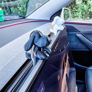 May include: Two dog-shaped car accessories, one black and one white, are perched on the dashboard of a car. The black dog is in the foreground, while the white dog is in the background. The car's interior is visible, with a screen and other details.
