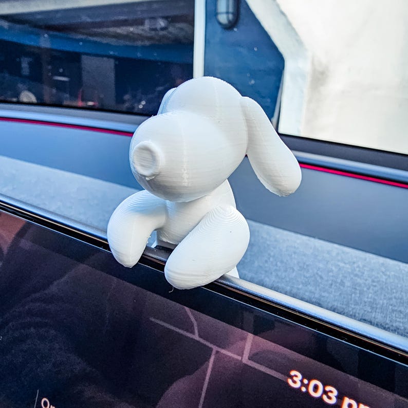K&ouml;nnte beinhalten: Eine wei&szlig;e, 3D-gedruckte Figur eines Cartoon-Hundes, platziert auf einer dunklen Oberfl&auml;che. Der Hund hat gro&szlig;e Ohren und einen runden K&ouml;rper. Die Uhrzeit 15:03 Uhr ist auf einem Bildschirm zu sehen.