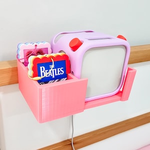 May include: A pink and purple wall-mounted shelf with a small pink and purple television-shaped box and a small red button on the side. The shelf is holding a few colorful toys, including a card with the text "The Beatles" on it.