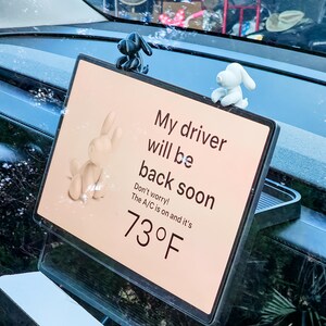 K&ouml;nnte beinhalten: Ein Schild mit dem Text "My driver will be back soon" und "73&deg;F" auf dem Armaturenbrett eines Autos. Das Schild ist mit kleinen Hunde- und Hasenfiguren verziert. Die Klimaanlage ist eingeschaltet.