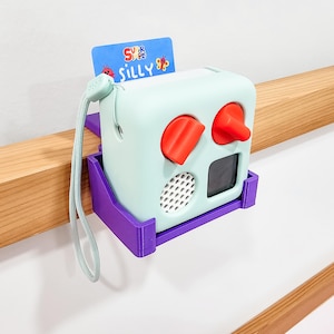 May include: A light blue toy with red buttons and a speaker grille. The toy is attached to a purple wall mount. The toy has a card on top that says "Super Simple Silly".