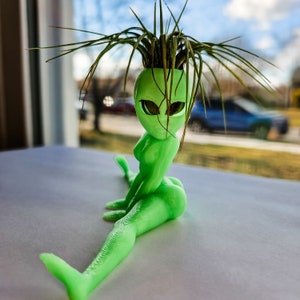 May include: A green alien figurine with large eyes, sitting in a split position with its legs spread apart. The figurine is made of plastic and has a plant growing out of its head.