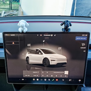 May include: A car dashboard with a large screen displaying a white car. Two dog figurines, one white and one black, sit on top of the screen. The screen shows various car controls and information.