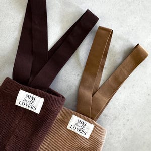 Könnte beinhalten: Nahaufnahme von zwei Cord-Latzhosen in Braun und Beige. Jede Hose hat ein weißes Etikett mit der Aufschrift "MINI MILK LOVERS". Die Latzhosen haben braune Träger und werden auf einer hellgrauen Oberfläche präsentiert.