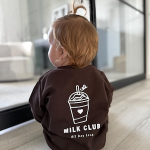 Könnte beinhalten: Ein braunes Sweatshirt mit einer weißen Grafik eines Getränkebechers und dem Schriftzug "MILK CLUB All Day Long". Das Sweatshirt wird von einem Baby mit hellbraunem Haar getragen.