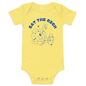 Könnte beinhalten: Gelber Baby-Body mit kurzen Ärmeln. Der blaue Text lautet "EAT THE RICH" über einer Cartoon-Blumenfigur, die ein Friedenszeichen gibt. Die Figur trägt Turnschuhe und hat ein lächelndes Gesicht.