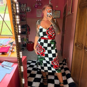 May include: A midi-length dress featuring a black and white checkered pattern with large red rose graphics and green leaves. The dress has thin shoulder straps, a V-neckline, and a side slit. A thin red belt with a heart-shaped buckle is worn at the waist.