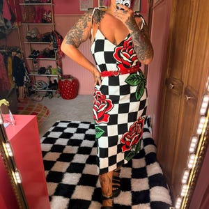 May include: A midi-length dress featuring a black and white checkered pattern with large red rose graphics and green leaves. The dress has thin shoulder straps, a V-neckline, and is cinched at the waist with a thin red belt featuring a heart-shaped buckle.