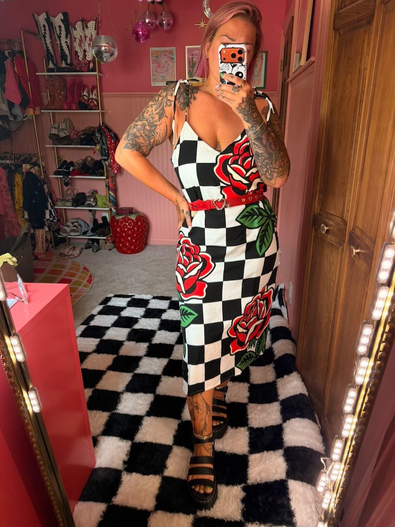 May include: A midi-length dress featuring a black and white checkered pattern with large red rose graphics and green leaves. The dress has thin shoulder straps, a V-neckline, and is cinched at the waist with a thin red belt featuring a heart-shaped buckle.