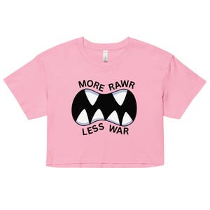 Puede incluir: Una camiseta corta rosa con el texto "MORE RAWR LESS WAR" sobre un gráfico de boca de dibujos animados con dientes blancos y un contorno negro. La camiseta tiene mangas cortas y cuello redondo.