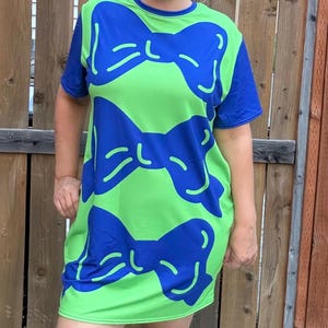 Peut inclure: Une robe t-shirt à manches courtes vert citron vif avec un motif répétitif de grands nœuds bleu roi audacieux. Le vêtement présente une encolure ronde et des manches bleu roi unies, avec l'imprimé de nœuds couvrant tout le panneau avant.
