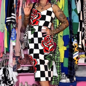 May include: A sleeveless midi dress featuring a black and white checkered pattern with large red rose graphics and green leaves. The dress has thin shoulder straps and a fitted silhouette. A small white tag hangs from the side of the garment.
