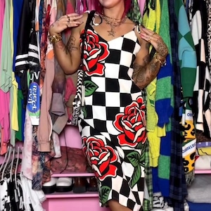 May include: A sleeveless midi dress featuring a black and white checkered pattern adorned with large red roses and green leaves. The dress has thin shoulder straps and a straight neckline, worn with red flat shoes and dark oval sunglasses.