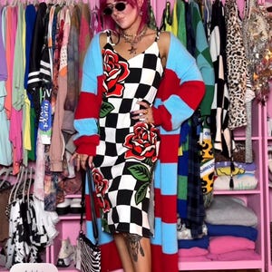 May include: A midi-length dress featuring a black and white checkered pattern with large red rose graphics, worn with a long-sleeved, open-front cardigan with horizontal red and light blue stripes, and a pair of red flat shoes with a strap.