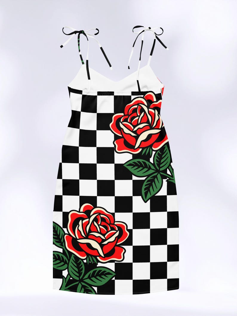 May include: A sleeveless dress featuring a black and white checkered pattern throughout. The garment is adorned with two large, vibrant red roses with green leaves and stems. It includes thin, adjustable tie-up shoulder straps at the top.